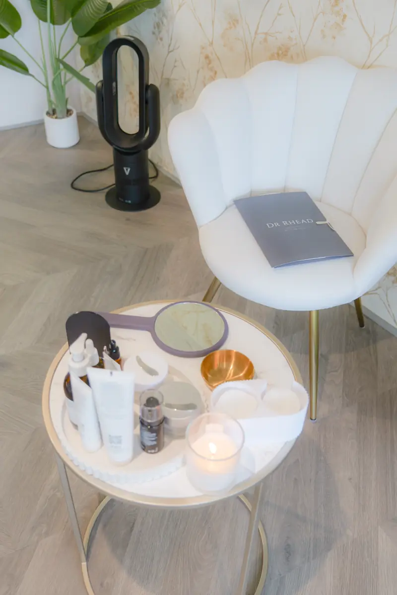 A scallop-backed white consultation chair with a Dr Rhead Aesthetics folder on the seat, beside a table of Medik8 skincare products and a hand mirror.