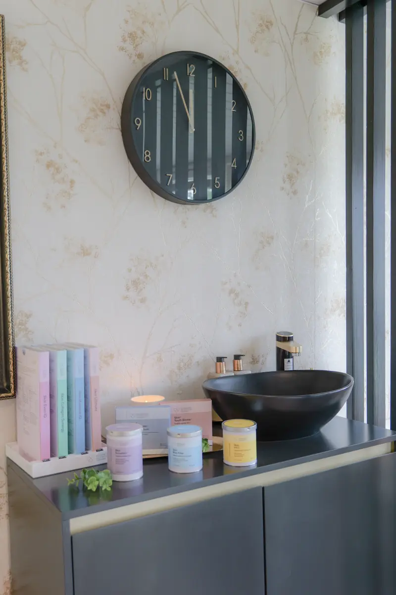 A treatment room corner with a black basin, candle, wall clock and pastel skincare jars on a dark shelf.